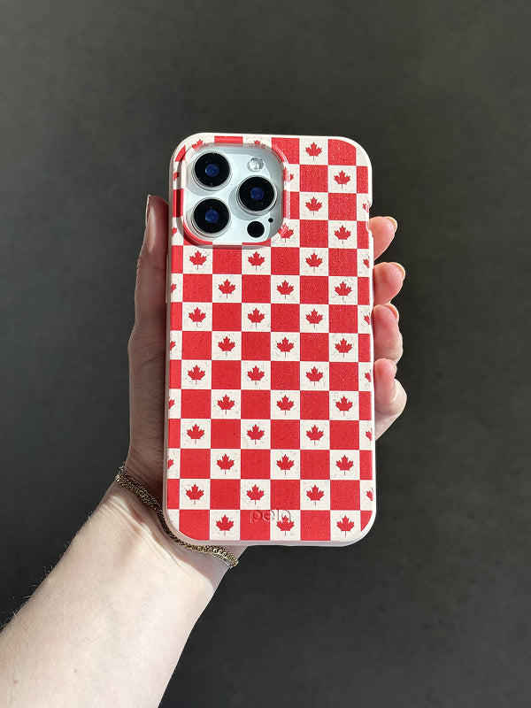 A hand holds a Pela phone case featuring a bold red-and-white checkerboard pattern with maple leaves in each square, inspired by the Canadian flag. The phone has a triple-lens camera, and the case is made from Pela’s compostable material, combining sustainability with patriotic style. The background is a neutral, softly lit surface.
