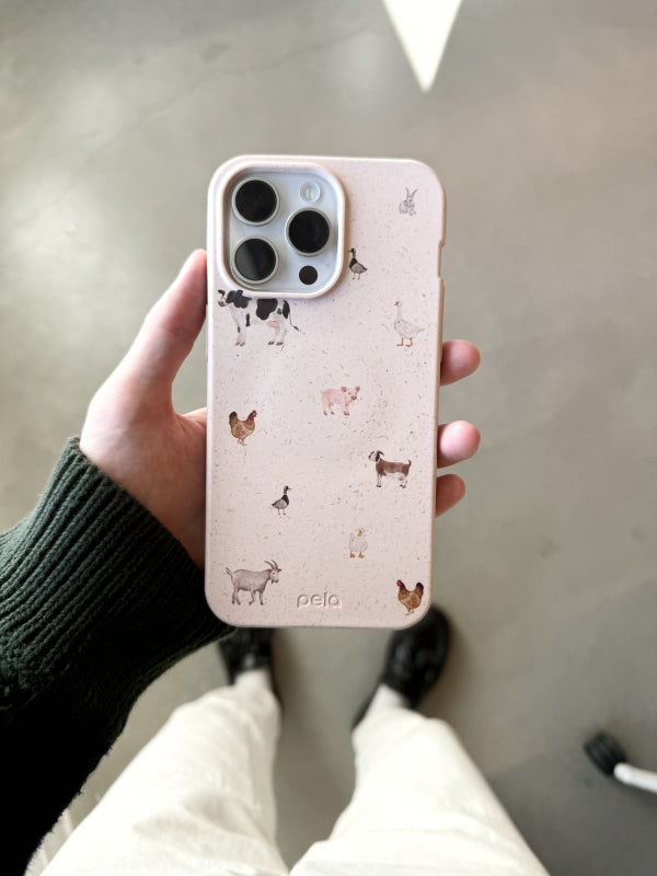 A hand holds the Pela Pasture Party phone case, featuring a whimsical design with various farm animals, including cows, chickens, goats, and ducks, scattered across a light background. The playful pattern brings a touch of rural charm, perfect for animal lovers and fans of countryside aesthetics.