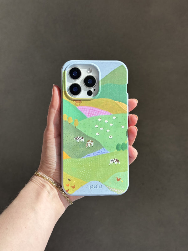 A hand holds a Pela phone case featuring the Farm Tales design. The case displays a colorful patchwork of rolling green hills, dotted with cows, sheep, chickens, and trees, creating a peaceful countryside scene. The phone has a triple-lens camera, and the case is made from Pela’s compostable material, blending sustainability with a charming, pastoral aesthetic. The background is a neutral, softly lit surface.