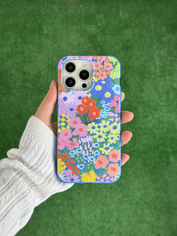 A hand holds a Pela phone case featuring the Painted Garden design. The case displays a vibrant, colorful floral pattern with a variety of illustrated flowers in shades of red, yellow, pink, blue, and green against a soft lavender background. Made from compostable materials, the case combines bold artistic style with eco-friendly function. The phone inside has a triple-lens camera, and the background is a grassy green surface.