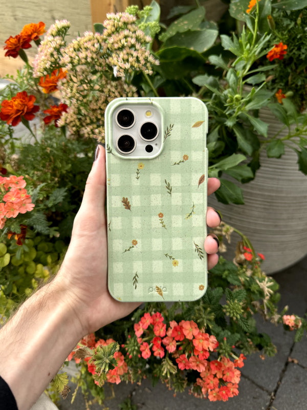 A hand holding the Pela Fall Gingham Phone Case against a backdrop of blooming autumn flowers. The phone case features a soft green gingham pattern with delicate fall-inspired elements like small leaves, acorns, and flowers scattered across the surface, evoking a cozy, seasonal vibe.