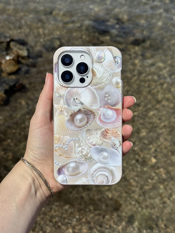 Hand holding a phone with the Sea Gems Phone Case by Pela, featuring a pearlescent seashell and pearl collage design, against a sparkling shoreline background.