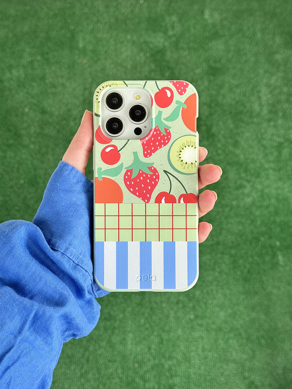 A hand holds a Pela phone case with the Fresh Picnic design, featuring a colorful collage of summer picnic elements like cherries, flowers, lemons, and playful patterns on a speckled background. The design is vibrant and eclectic, capturing a fun, seasonal vibe. The phone has a triple-lens camera, and the background is a bright green grassy surface.







