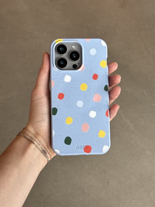 Hand holding a phone with the Rainbow Dots Case, featuring a pastel blue background and scattered multicolored polka dots, against a neutral brown surface.
