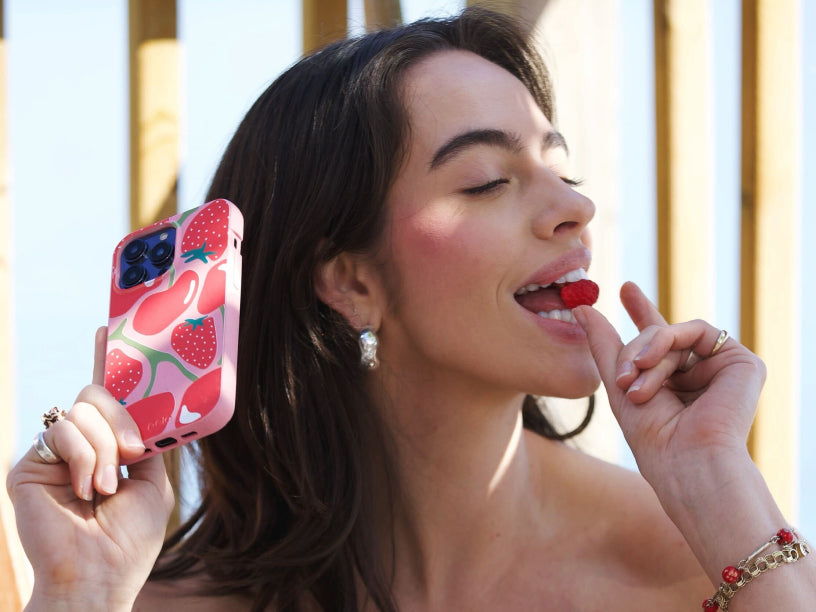 A close-up of the Pela Cherry Berry Phone Case, showcasing a playful design of cherries and strawberries scattered across a soft pink background. The phone case is being held in hand, offering a vibrant and fruity aesthetic perfect for summer.