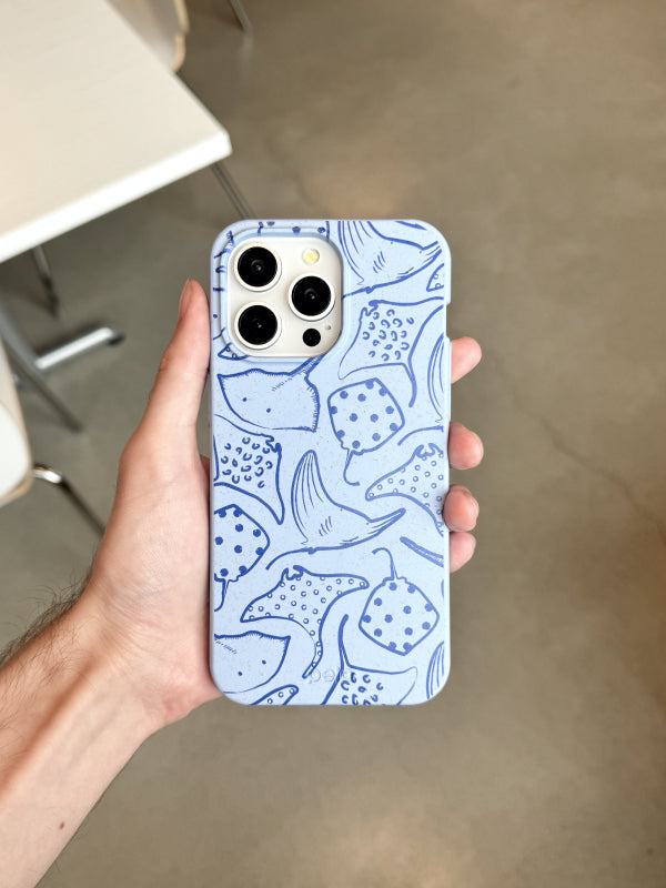 Hand holding Pela’s Stingray Fever Phone Case, featuring a playful pattern of blue stingrays on a light blue background. The earth-friendly case highlights the beauty of sea life with its intricate, hand-drawn stingray illustrations.