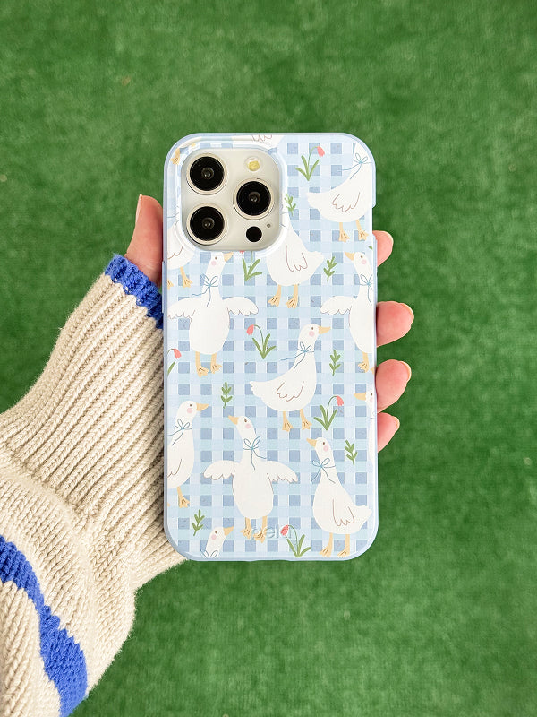 A hand holds a Pela phone case with a Blooming Quackers design, featuring white ducks with blue bows on a soft blue gingham background. Scattered tulips and greenery add a springtime feel. The phone inside has a triple-lens camera, and the background is a bright green grass-like surface.
