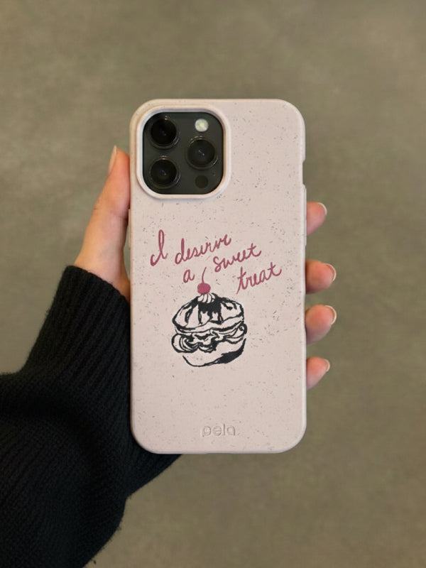 Person wearing black knit sweater holds iPhone with cream Pela phone case featuring red script text reading 'I deserve sweet as a treat' and black line drawing of cream puff with cherry on top, against neutral beige background