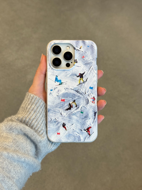 Person wearing grey knit sweater holds iPhone with Pela Painted Slopes phone case featuring white ski slopes with colorful skiers and snowboarders carving through powder, gray background